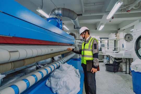 Confident Asian maintenance technician with a digital tablet checking linen quality on a large industrial flatwork ironer in a smart commercial laundry facility for hospital hygiene.