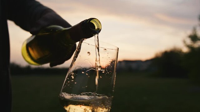 Grapes transform into a fine beverage as a hand pours white wine into a glass, splashing brightly against the backdrop of a stunning sunset.