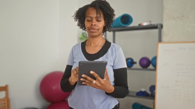 Woman standing and tapping a tablet with her hands while checking a workout plan near yoga mat, exercise balls and whiteboard in a studio; focus wellness.