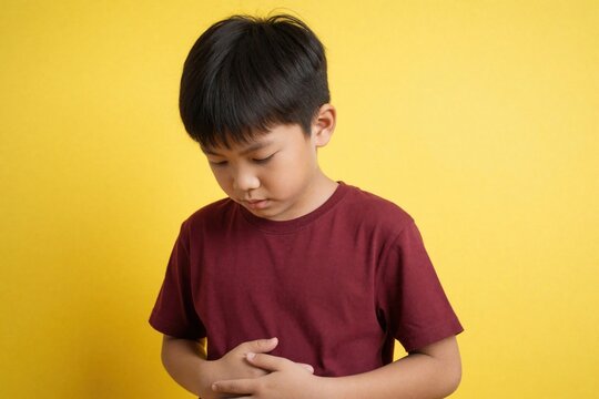 Young Asian boy holds stomach in discomfort against bright yellow background. Looks down with pained expression, indicating stomach ache illness. Image suitable for health-related topics,