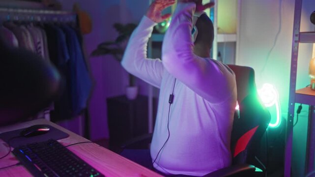 Man lifting headset with one hand while holding a wireless game controller and resting near a keyboard and mouse at a neon-lit desk with shelving and clothing rack in a studio; late night fatigue.