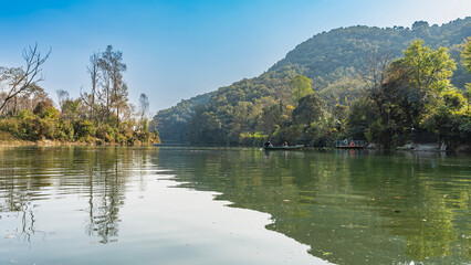 Fototapeta premium Beautiful calm lake. There is green vegetation on the shores. The tourist boat is far away. A mountain against a blue sky. Reflection on the water. Nepal. Pokhara. Phewa lake.