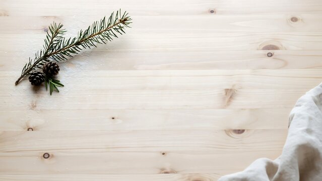 A single green pine branch with two small brown pine cones resting on a light wooden surface with a white fabric cloth in the corner.