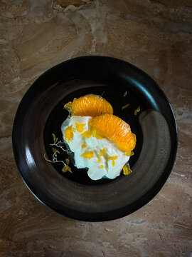 A high-angle studio shot of thick creamy greek yogurt decorated with fresh orange slices and citrus zest, elegantly served on a minimalist black ceramic plate with copy space.