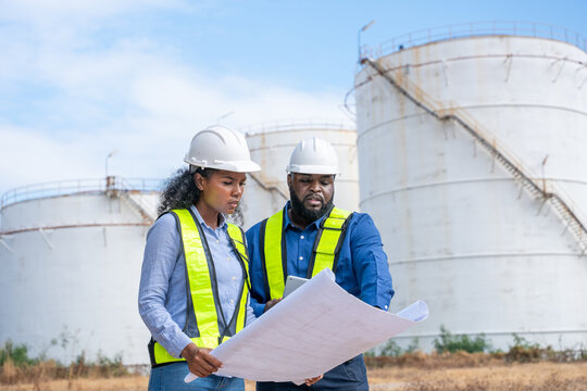 Engineers wearing safety gear, including hard hats examining survey a large blueprint tablet standing industrial facility gas or oil refinery engaged in a job requires high safety standards concept.	