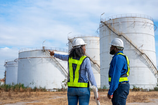 Engineers wearing safety gear, including hard hats examining survey a large blueprint tablet standing industrial facility gas or oil refinery engaged in a job requires high safety standards concept.	