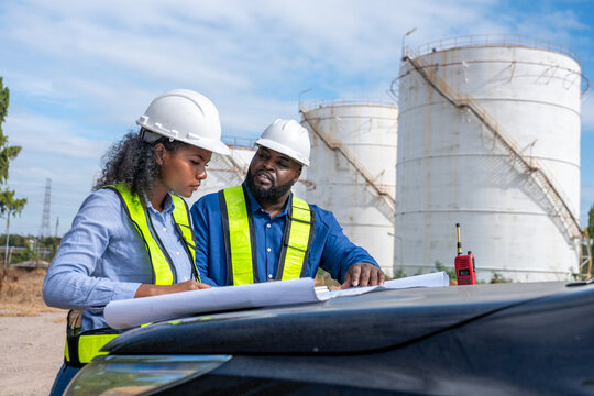 Engineers wearing safety gear, including hard hats examining survey a large blueprint tablet standing industrial facility gas or oil refinery engaged in a job requires high safety standards concept.	