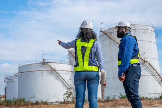 Engineers wearing safety gear, including hard hats examining survey a large blueprint tablet standing industrial facility gas or oil refinery engaged in a job requires high safety standards concept.	