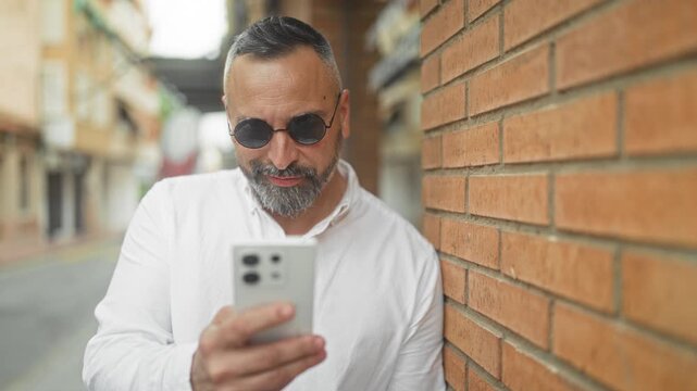 Man holding smartphone and scrolling with thumb while leaning on brick wall street, wearing round sunglasses and white shirt; calm focus.