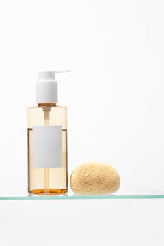Clear pump bottle filled with golden liquid next to natural sponge on glass shelf. Minimalist spa and beauty concept for luxury skin cleansing.