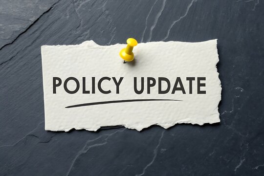 Policy update text written on a torn piece of white paper attached to a dark slate background with a yellow pin