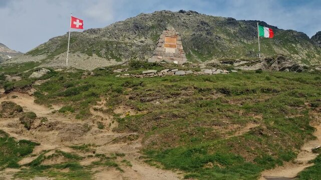 Border point at Spluga Pass showing the Italian and Swiss flags gently waving in the wind at high altitude, surrounded by a rugged alpine landscape marking the mountain border between the countries