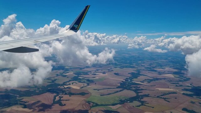 Passenger window view from an airplane crossing Paran&aacute;, Brazil, with expansive farmland, rural landscapes, and agricultural patterns visible below through scattered clouds during the flight
