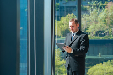 Fototapeta premium Businessman using tablet standing beside office window. Professional reviewing business information, checking reports, managing projects and working with digital technology in corporate workspace.