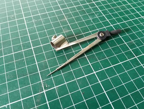Metal drawing compass with a sharp needle point and pencil holder, used for drawing circles and arcs in technical drawing, drafting, geometry, and school work, isolated on a green cutting mat.