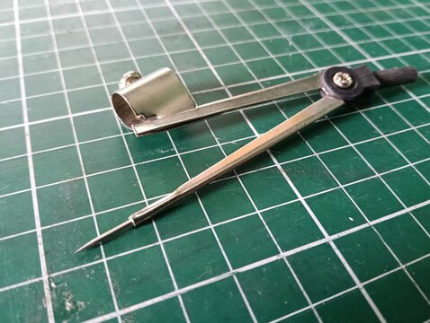 Metal drawing compass with a sharp needle point and pencil holder, used for drawing circles and arcs in technical drawing, drafting, geometry, and school work, isolated on a green cutting mat.