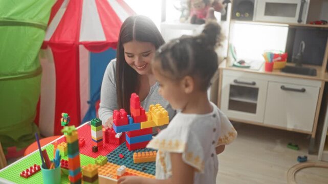 Woman helping little girl stack colorful plastic blocks with hand on table near a play tent and toy kitchen in building; early learning joy.