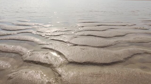 A wide-angle landscape shot of natural wavy sand ripples on a coastal mudflat beach during low tide, reflecting bright sunlight against a soft cloudy sky at the horizon.