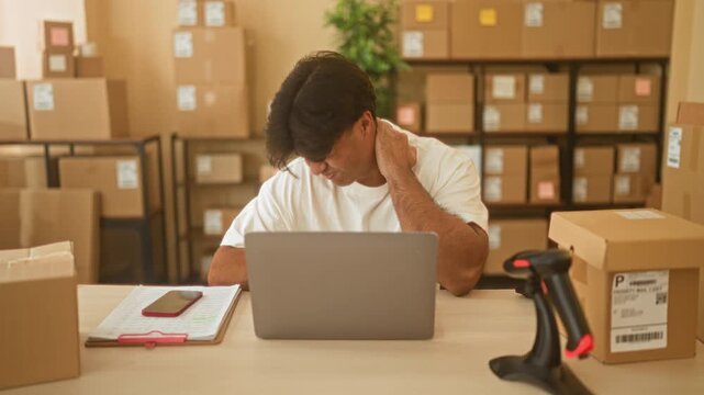 Man rubbing neck while checking laptop and parcel list amid stacked boxes and barcode scanner in a building fulfillment area; stress fatigue.