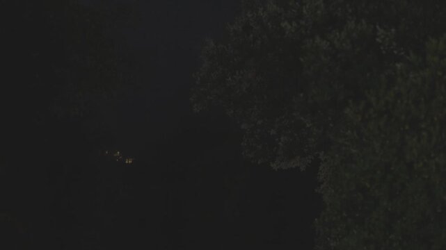 A rural house with glowing windows stands in a dark forest at night during a rain storm with lightning flashes illuminating the sky and trees