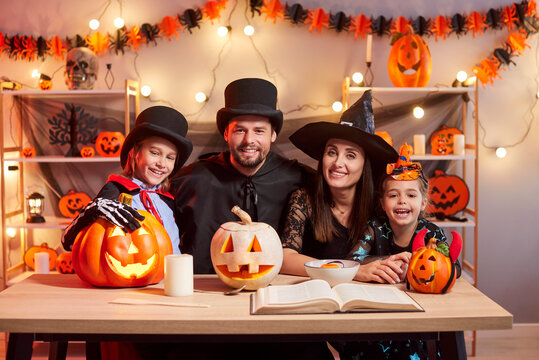 Portrait of smiling happy young family of mother, father and children wearing masquerade costumes celebrating Halloween party at home sitting at festive table with pumpkins decorations