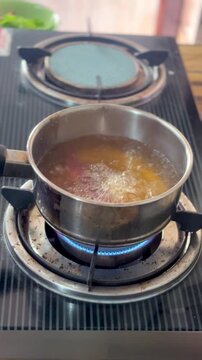 Boiling liquid bubbles inside small stainless steel saucepan heating over gas stove burner flame, preparing warm soup inside domestic home kitchen counter space, nobody present.