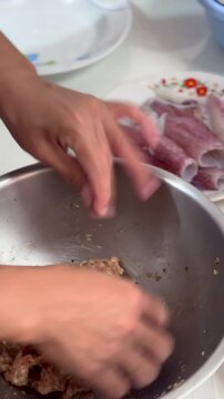 Home cook hand mixes seasoned minced pork stuffing inside stainless steel bowl, preparing raw squid seafood ingredients nearby inside kitchen workspace, making traditional Asian stuffed calamari.