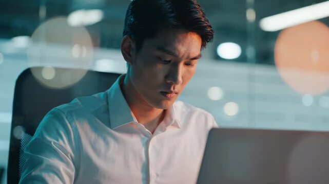 Focused young man working intently on his laptop in a modern office setting, illuminated by soft lighting, conveying concentration and determination.