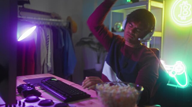Man at desk typing on keyboard, removing white gaming headset with one hand, popcorn bowl and controller on desk, neon sign on shelf in studio; concentration.