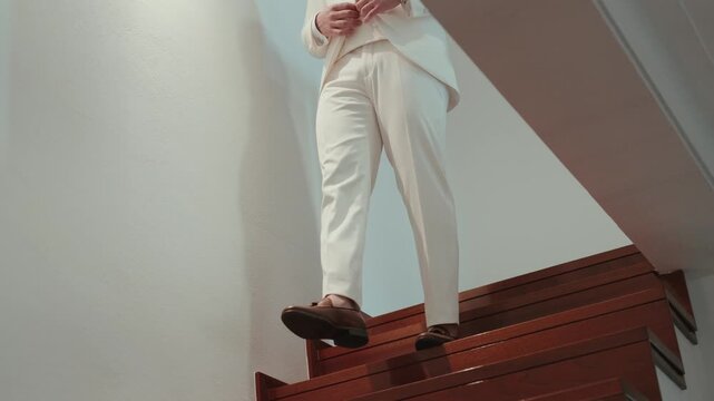 Groom in cream trousers and loafers descending wooden stairs indoors