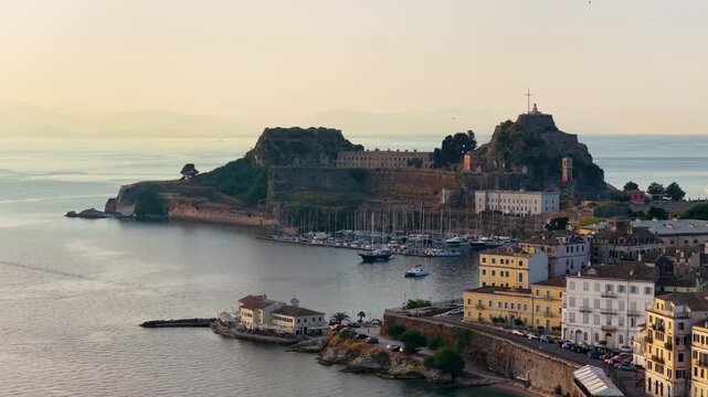 A stunning wide shot of the historic Old Fortress (Palaio Frourio) in Corfu, Greece, during a golden sunrise. The calm sea surrounds the fortress, with yachts docked in the marina and birds flying acr