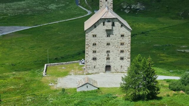 Historic Old Hospice (Alter Spittel) on Switzerland's Simplon Pass stands amid lush green meadows and stunning alpine scenery on a bright, sunny summer day