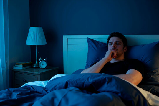 Young adult Caucasian man lying awake in bed, thinking during nighttime insomnia and sleep struggle. Useful for sleep disorder, stress, mental health, fatigue campaigns