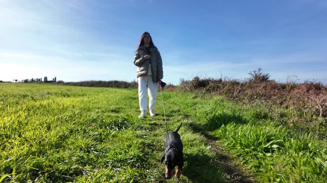 Woman walking a dachshund toward the camera on a green countryside trail
