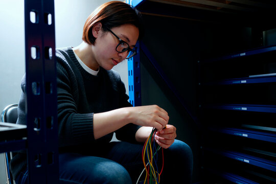 Asian young adult woman connecting network cables in server rack, managing IT infrastructure and hardware setup. Ideal for technology support, data center, cybersecurity marketing