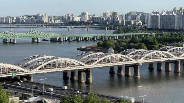 Clear day timelapse view overlooking the Han River, Nodeul Island, Hangang Bridge, and Hangang Railway Bridge with a train crossing, featuring moving traffic on Olympic-daero and apartments in Yongsan