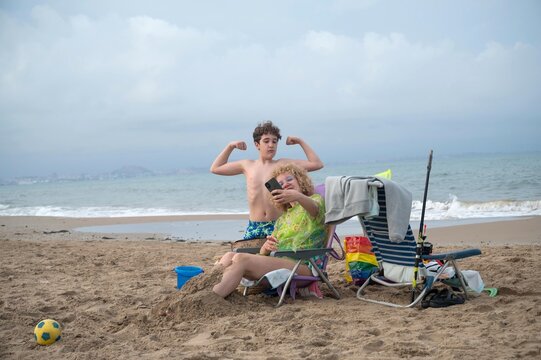 Mother taking a selfie with her son striking a playful muscle pose on a quiet sandy beach during a cloudy summer day, capturing fun family vacation memories by the sea