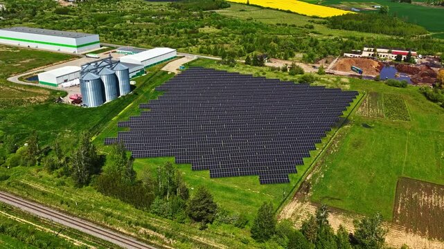 Aerial view of solar farm with rows of photovoltaic panels next to grain silos and industrial buildings in a rural landscape in Riga, Latvia.