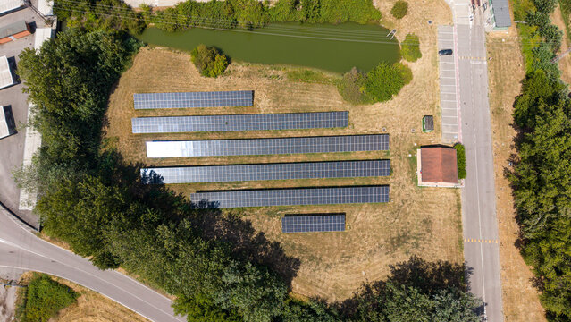 Aerial view of a solar farm with rows of photovoltaic panels, a small building, and a pond surrounded by trees and a road in Tavazzano con Villavesco, Lombardy, Italy.