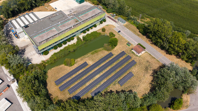 Aerial view of a solar farm featuring rows of photovoltaic panels, an industrial building, a small pond, and surrounding greenery in Tavazzano con Villavesco, Lombardy, Italy.
