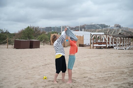 Mother helping her son put on a sweatshirt after playing on the beach during a cloudy summer day, sharing a caring family moment by the sea after outdoor activities