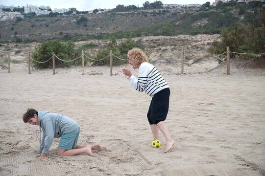 Mother cheering and encouraging her son while they play together on a sandy beach during a cloudy summer day, sharing a joyful and active family vacation by the sea