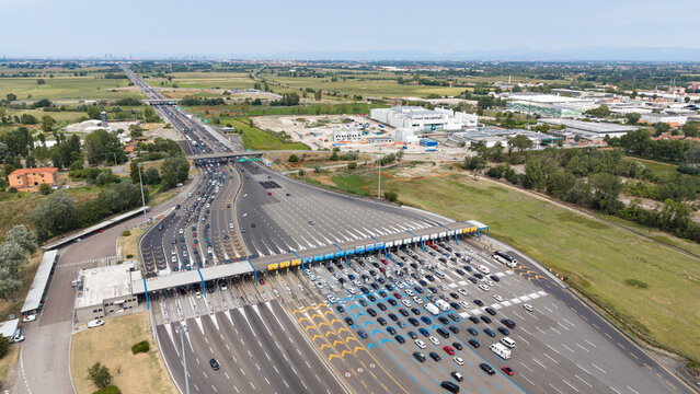 Aerial view of Milano Sud toll plaza on A1 motorway with heavy vehicle traffic, highway infrastructure, and surrounding landscape in Melegnano, Lombardy, Italy.