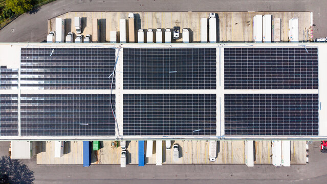 Aerial view of a logistics hub with solar panels on the warehouse roof and freight trucks parked at loading docks in Melegnano, Lombardy, Italy.