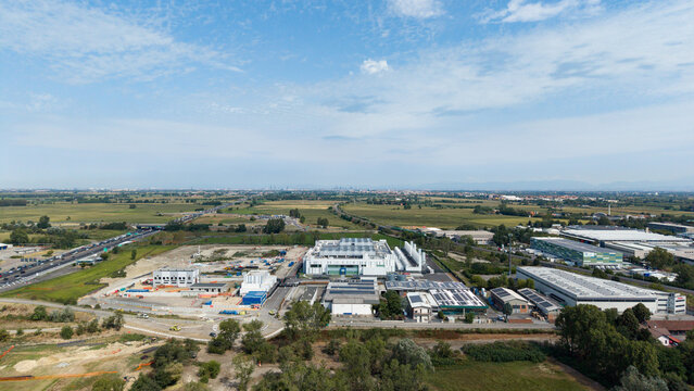 Aerial view of a modern data center campus with industrial buildings, solar panels, and surrounding green fields near a highway in Melegnano, Lombardy, Italy.