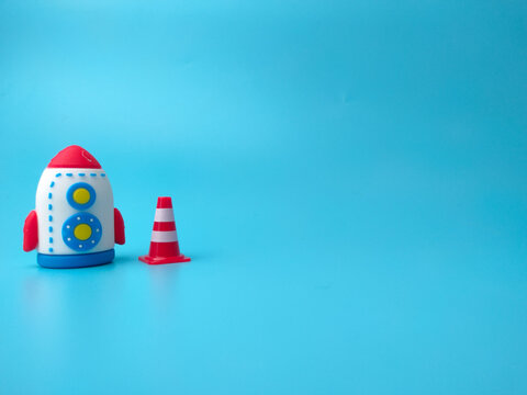 A toy rocket stands alongside a miniature red and white traffic cone against a solid blue backdrop