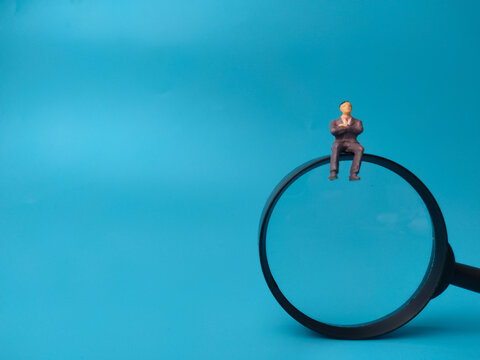 A miniature figurine of a businessman sits atop the rim of a magnifying glass on a blue background