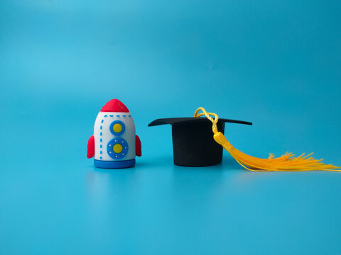 A toy rocket stands next to a miniature black graduation cap with a yellow tassel over blue background