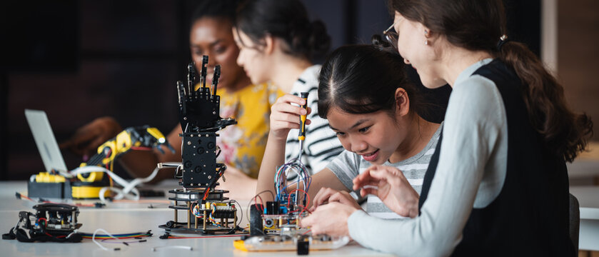 STEM Education with an Asian student building a wheeled robot as a female teacher offers practical guidance beside robotic equipment in a technology class