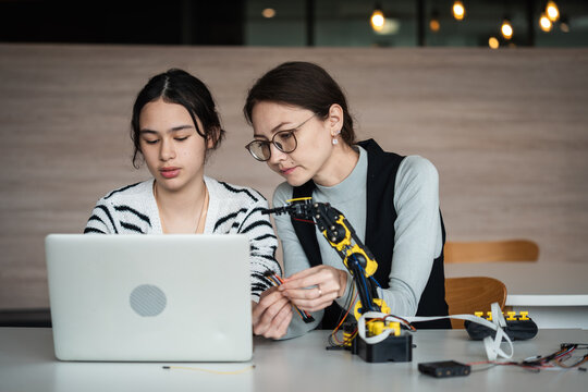 Robotics Education with an Asian female teacher guiding a young woman using a laptop beside a robotic arm during a practical technology lesson in class